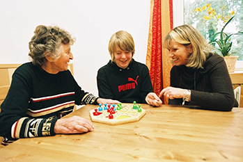 pflege-daheim-muenchen-formular-02 3 Personen sitzen am Tisch und spielen ein Brettspiel. Alle lächeln.
