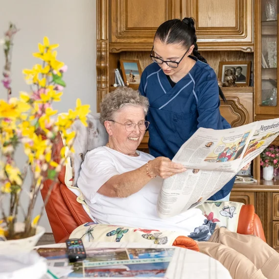pflegedienst-daheim-ambulante-pflege-muenchen-02 Pflegekraft steht im Wohnzimmer einer Klientin und am Sessel und schaut zu ihr hin. Klientin schaut lächelnd im Sessel sitzend in eine Zeitung.