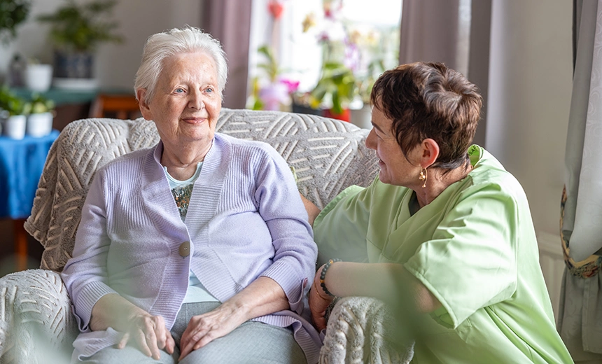 Klientin sitzt im Wohnzimmer auf einem Sessel und lächelt zu einer weiblichen Pflegekraft, die sich auf der Sessellehne auflehnt und zur Klientin schaut.